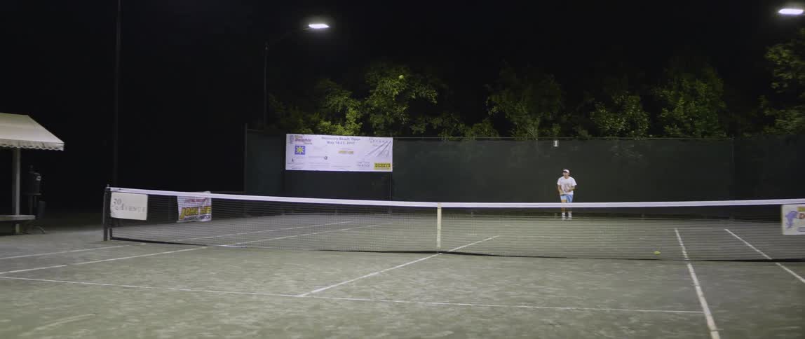 Two men play tennis on an outdoor court at night