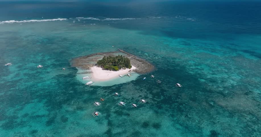 Boats float in the blue waters near a small island with a white sand beach during the daytime