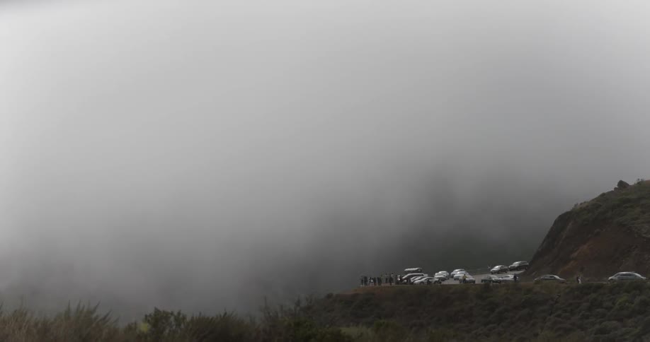 People and cars gather near a mountain lookout shrouded in fog