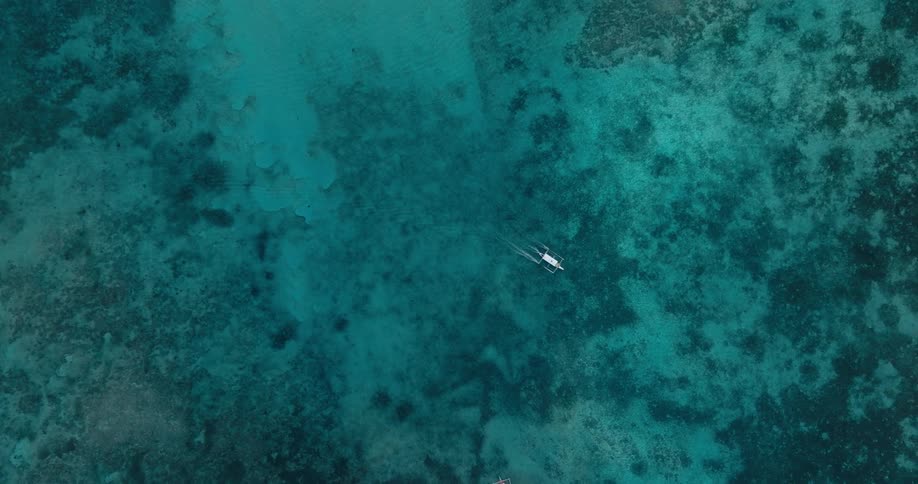 An aerial view shows a small boat navigating the clear turquoise waters revealing the coral reef patterns beneath the surface