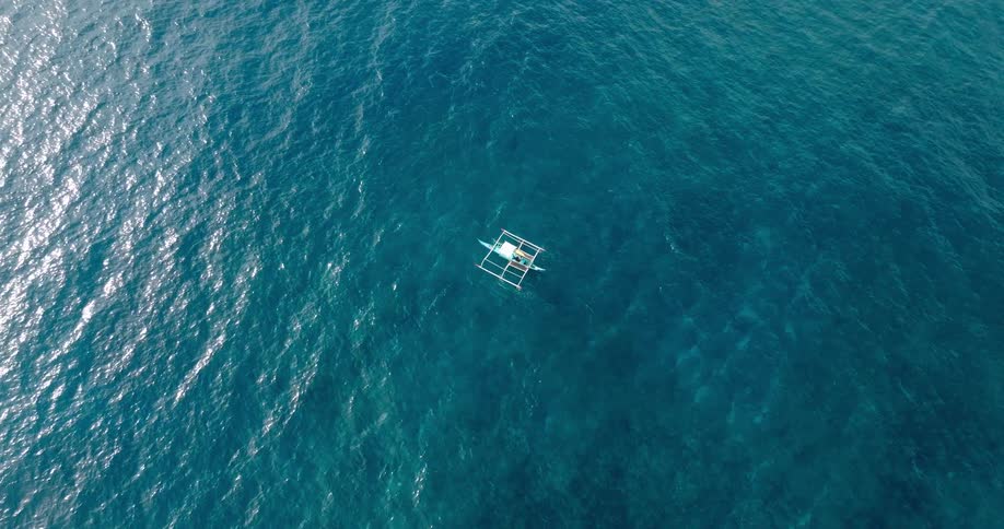 An aerial view of a small white boat with outriggers floating in the vast deep blue expanse of the ocean