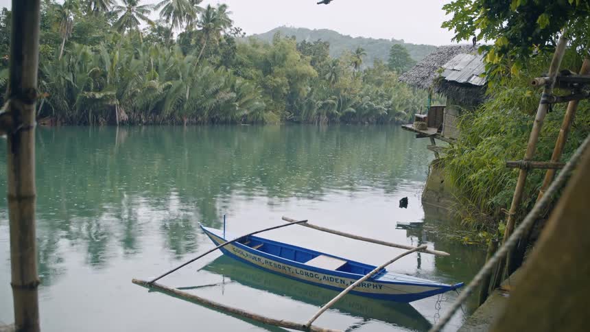 From a riverside hut a boat floats calmly on the water as gentle rain creates ripples across the surface surrounded by lush tropical vegetation