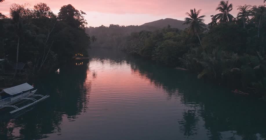 A tranquil river scene at dusk framed by lush jungle foliage and reflecting a vibrant pink sky