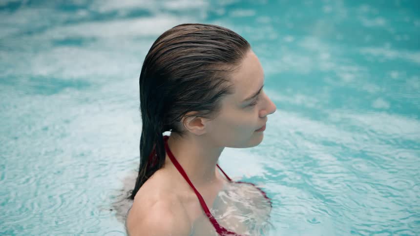 A young woman in a red bikini top with wet slicked back dark hair is in a swimming pool with clear blue water that ripples around her as she moves her left arm to and fro looking up to her right at someone and giving a slight content smile