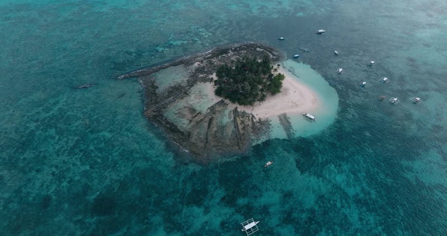 An aerial view flies lower toward a small tropical island fringed with palm trees and surrounded by turquoise water as boats dock nearby and people walk along the sandy beaches