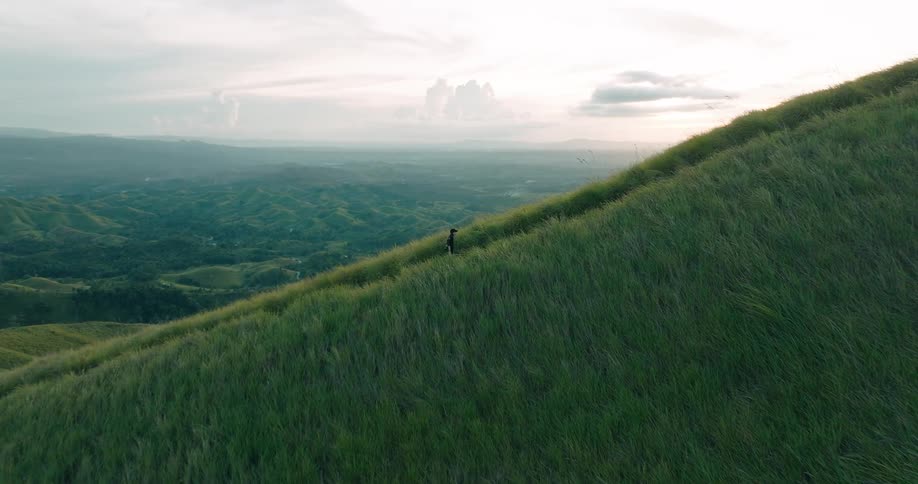 A lone figure walks along a lush green hillside overlooking a vast mountainous landscape under a cloudy sky