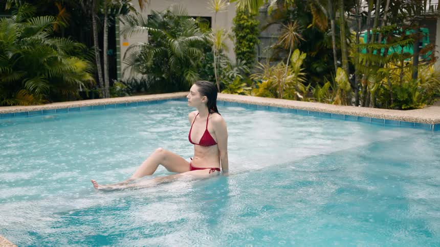 A woman in a red bikini sits in a shallow blue swimming pool surrounded by lush tropical foliage as it gently rains