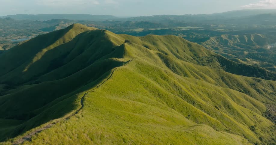 Aerial view captures rolling green hills stretching into the horizon bathed in warm sunlight under a partly cloudy sky
