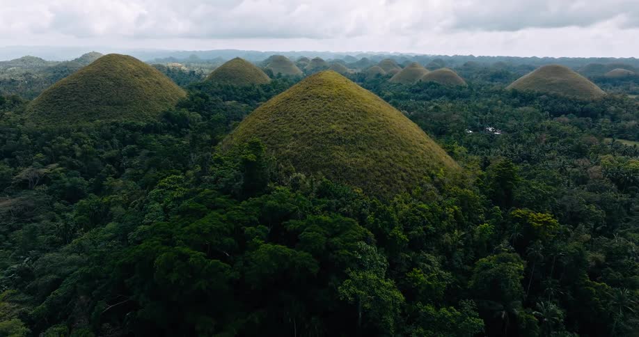 The aerial camera glides forward over the iconic chocolate hills surrounded by thick tropical jungle and layered under an overcast sky
