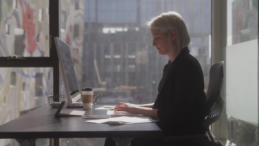 A woman with dark blazer and short light colored hair works at a computer in an office setting with sunlight streaming through a large window behind her