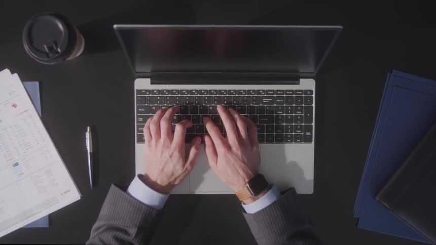 A man wearing a watch is typing on a laptop indoors