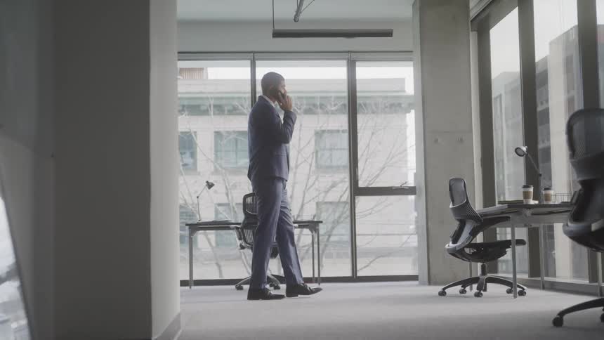 The man is walking across the office past a desk and chair while talking on his cell phone