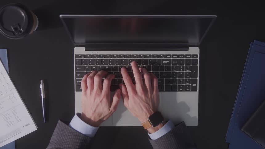 A man in a business suit types on a macbook at a dark desk viewed from above with coffee documents and folders beside him