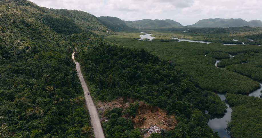 An aerial view of a winding road cutting through a lush green landscape of dense forest and meandering rivers framed by rolling hills under a cloudy sky with a biker riding a bike along the road