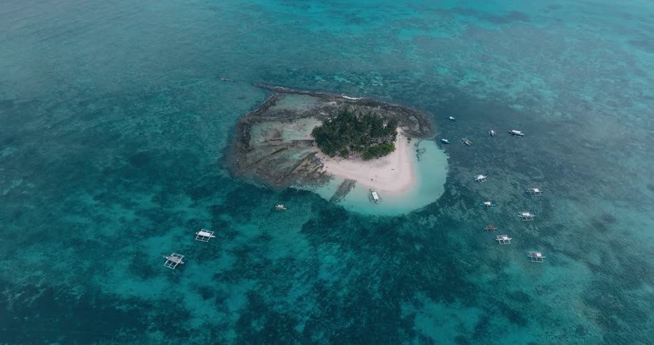 An aerial view shows a small lush island surrounded by vibrant turquoise waters dotted with numerous small boats