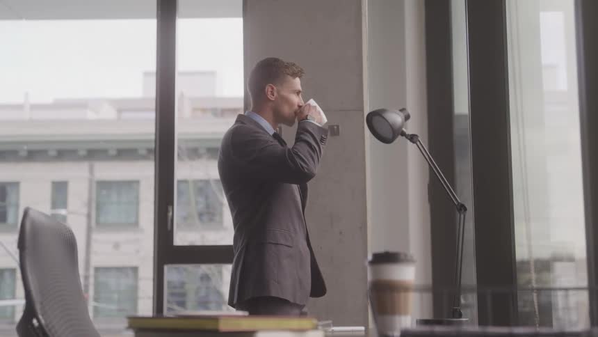 Professional male standing looking out office window drinking coffee