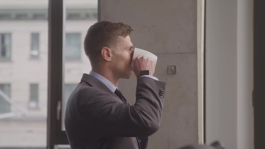 Businessman smiles and drinks coffee while looking out the office window