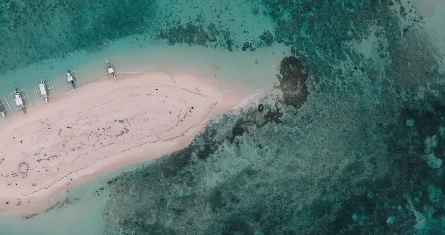 An aerial view captures a narrow strip of sandy beach bordered by clear turquoise water with small boats anchored nearby and a faint watermark visible on the sand