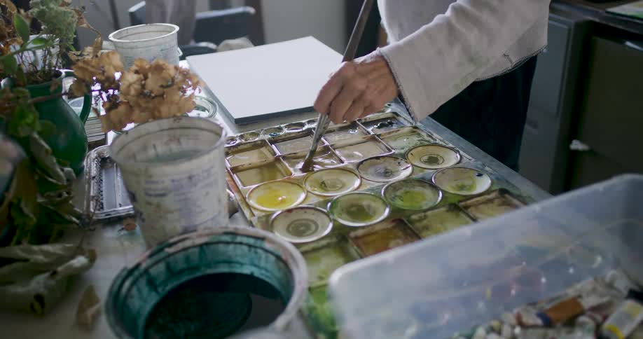 A hand holding a brush dips into a tray of various colored liquids surrounded by other art supplies