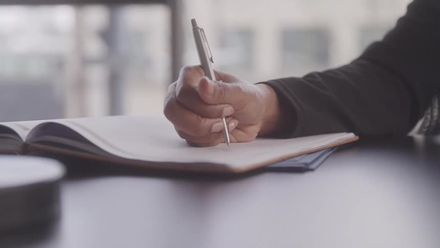 Close up of a person in a long sleeve shirt meticulously writing with a pen on a large open book placed on a desk