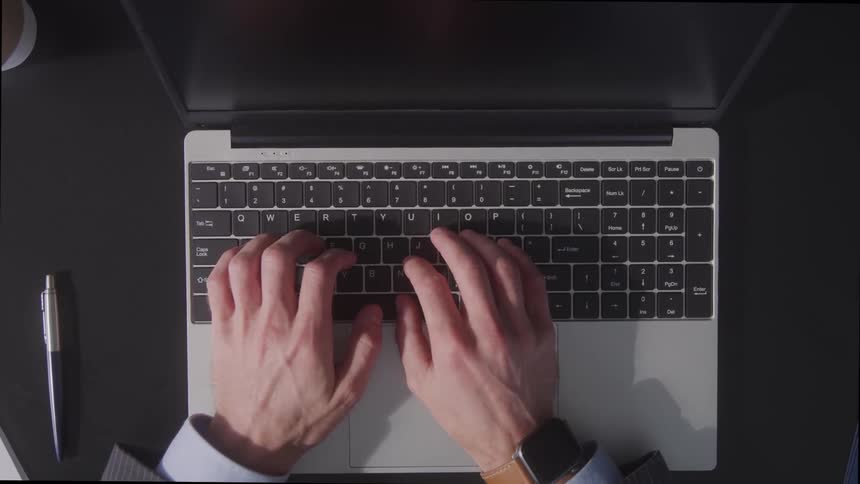 A mans hands typing on a laptop keyboard from an overhead perspective with a pen visible to the left and a watch on their wrist