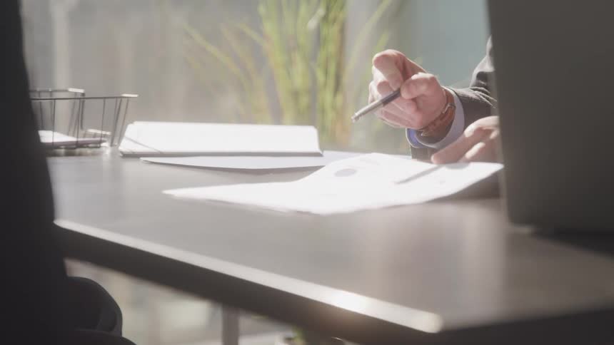 A businessman wearing a suit jacket is shown from the torso up sitting at a dark desk and using a pen to write on a piece of white paper