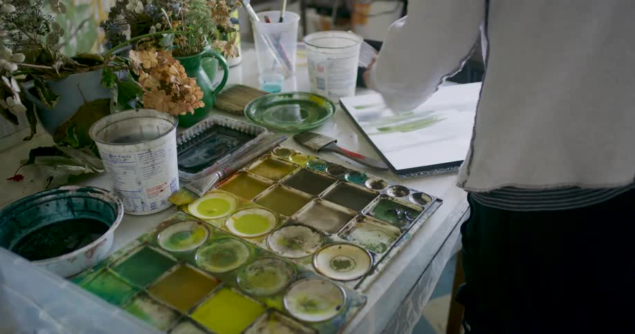 An artist works at a table covered with watercolor paints in a palette brushes water containers and a sketchbook showing painted strokes