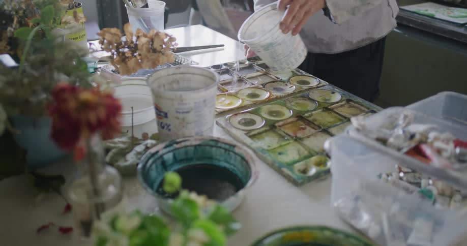 An artist carefully pours water into a large palette on a table filled with various containers plants and scattered art supplies