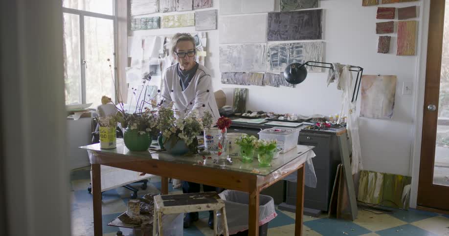 An artist in a workspace filled with paintings plants and art supplies working near a table showcasing vibrant floral arrangements