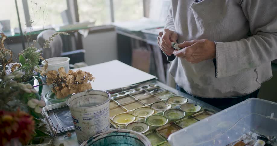 An artist carefully mixes colors on a large palette set on a table cluttered with containers plants and scattered art supplies