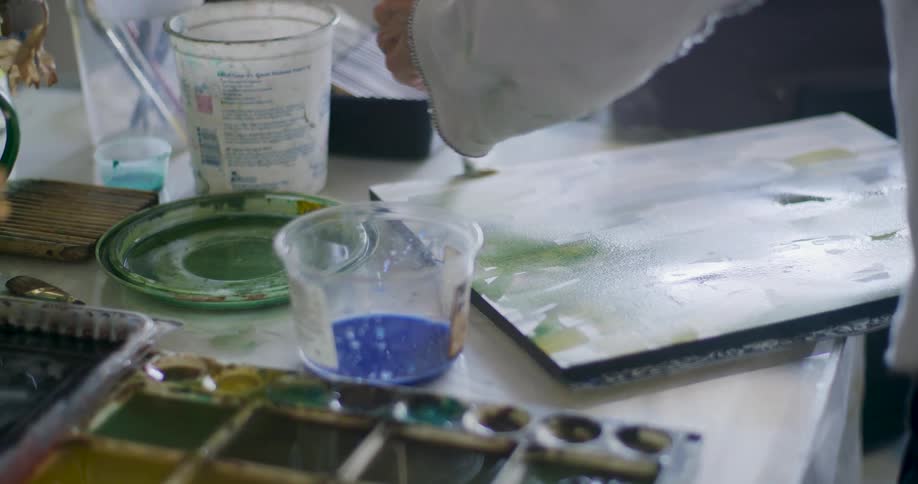 An artists workspace featuring a watercolor palette with various colors a clear cup containing blue water a painting in progress and other art supplies scattered on a light colored surface