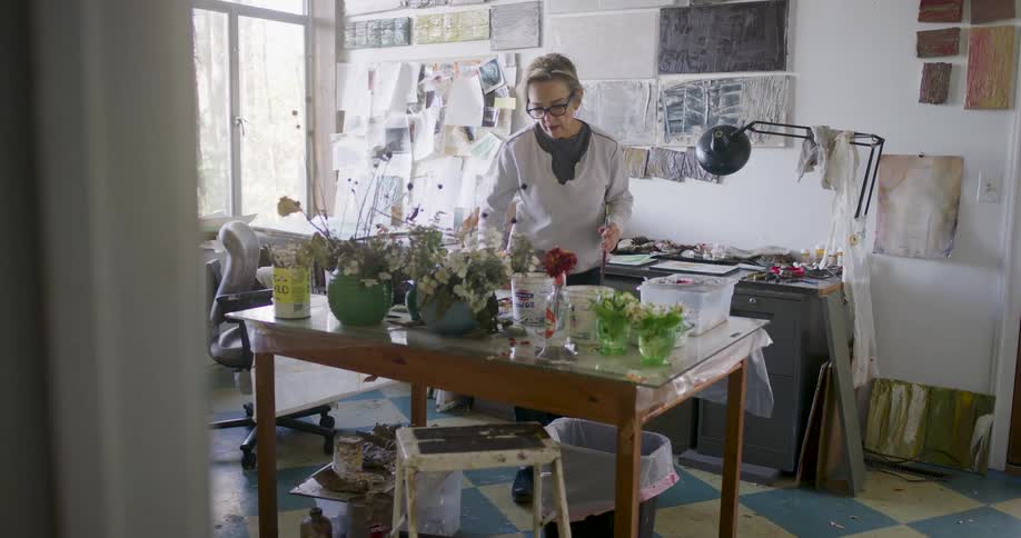 An artist stands in a cluttered studio space with tables holding various supplies and artwork on the walls