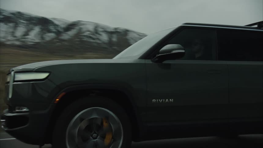 A darkcolored rivian r1s suv drives along a paved road with a yellow line against a backdrop of overcast skies and distant muted mountain