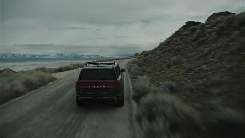 A dark rivian r1s suv with a roof rack is driving on a winding coastal road under a cloudy sky