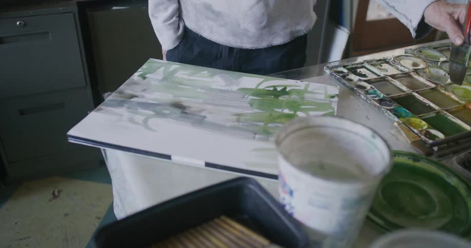 A man in a white shirt and dark pants paints on a canvas with a brush amidst a cluttered workspace with paints brushes and other art supplies