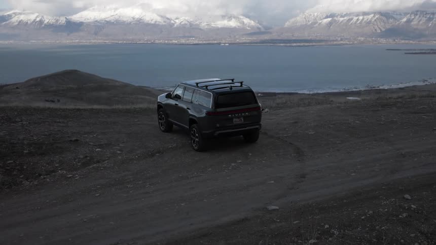 A dark colored rivian r1s suv is parked on a dirt road overlooking a lake and snow capped mountains under a cloudy sky