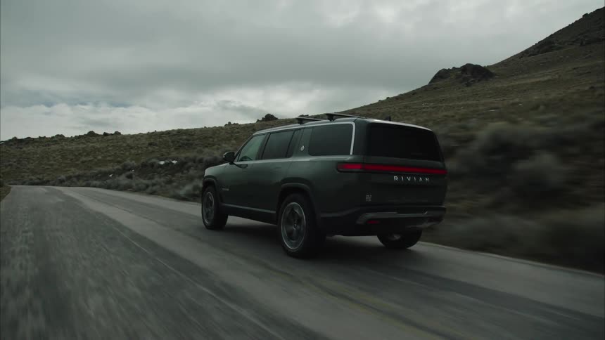 A dark colored rivian r1s suv drives on a paved road through a desolate mountainous landscape under an overcast sky
