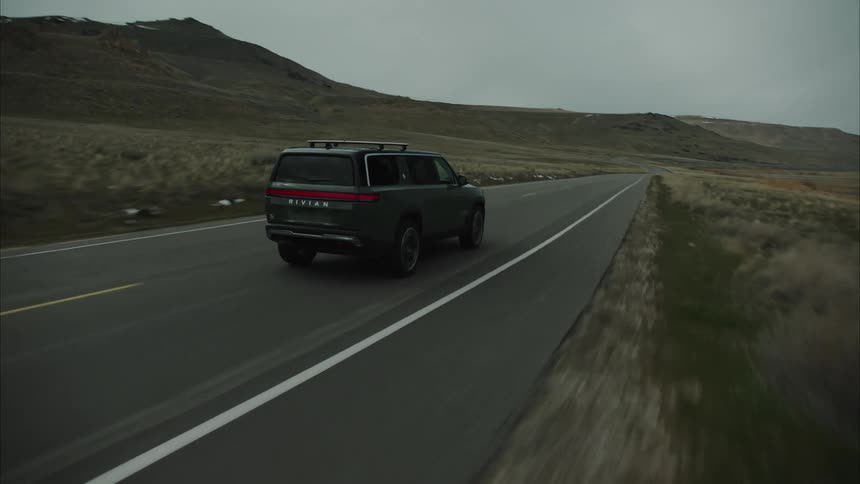 A dark colored rivian r1s suv driving on a paved road through a desolate mountainous landscape under an overcast sky