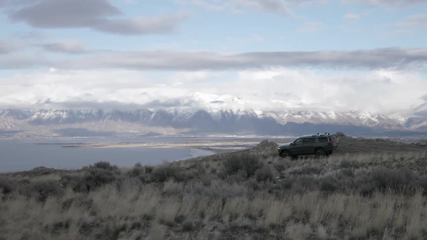 Sports utility vehicle travels along a coastal highway across the water from snowy mountains on a cloudy day