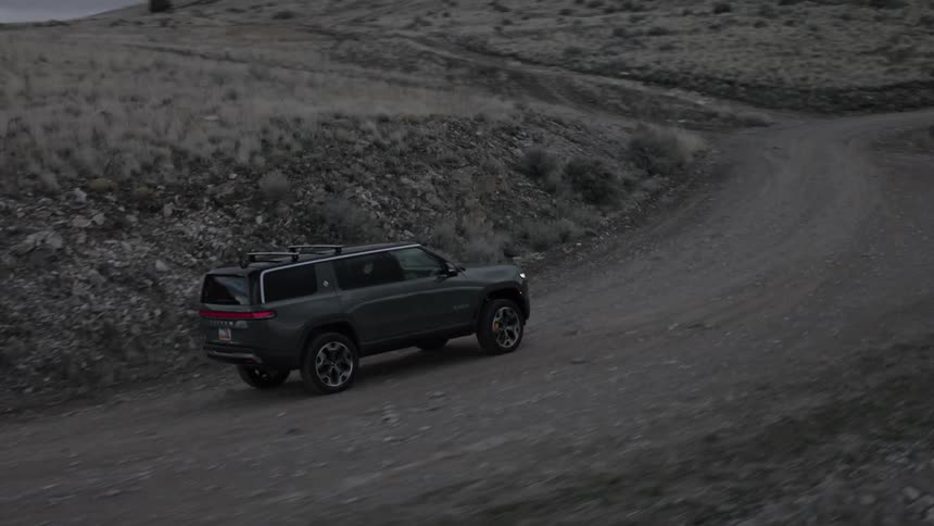 A dark colored rivian r1s suv driving up a rough rocky path on a hillside