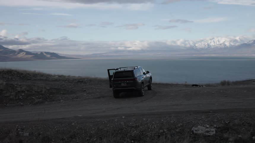 A person walks along a dirt road towards a parked vehicle in a scenic landscape featuring mountains and water