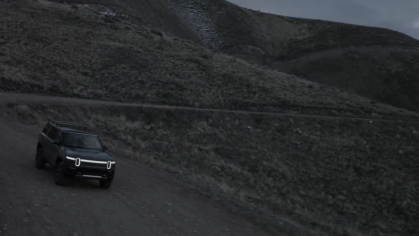A dark colored rivian r1s suv driving on a dirt road through a dark hilly landscape