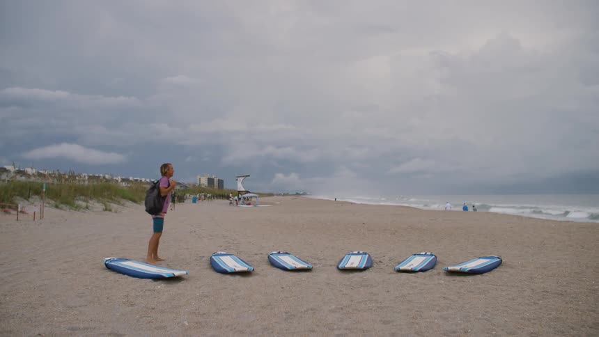 A man in shorts a pink shirt and wearing a backpack has set out six surfboards on the sand of a mostly empty beach under a grey cloudy sky before taking off his backpack and putting it on the sand