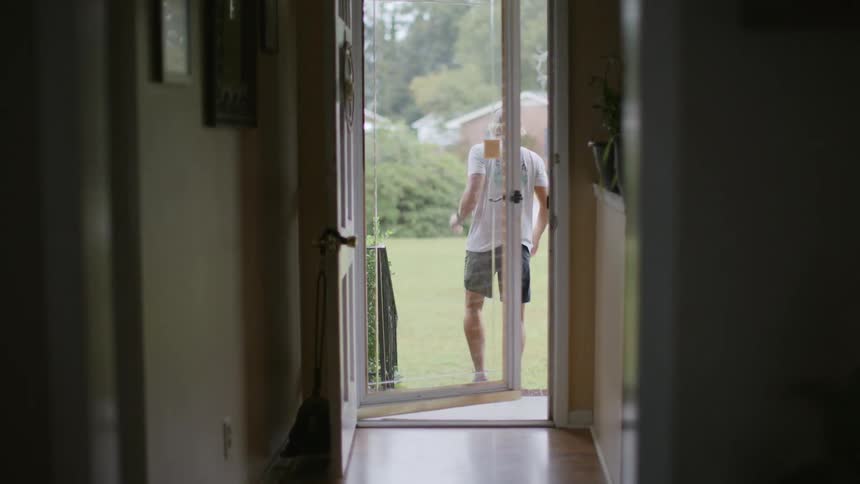 A man in shorts steps out of a front glass door into the sunlit yard likely heading out for a morning jog