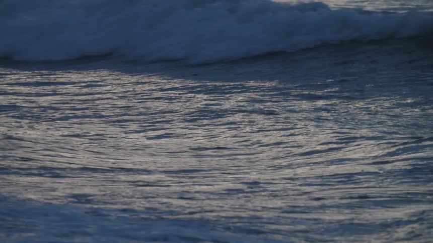 Waves gently crash along the shoreline the surface shimmering with reflections in the soft evening light