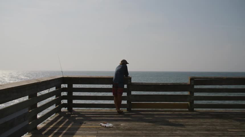 Man in a baseball cap standing at the end of a pier with a fishing pole nearby