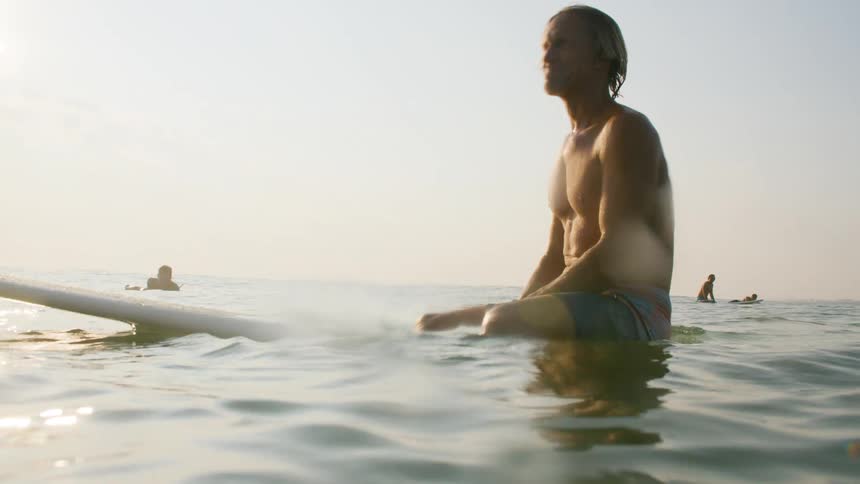 An older man with a serene smile sits knee deep in the ocean surrounded by other surfers