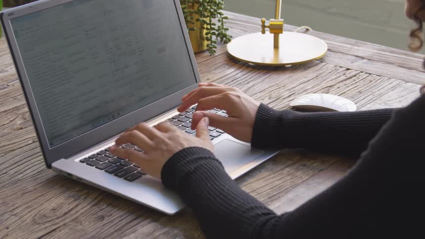 A woman types on a laptop at a wooden desk with a mouse and a small potted plant visible nearby
