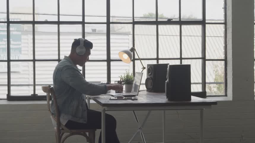 A man sits at a desk with a laptop working near a large window in what appears to be a modern office or studio space
