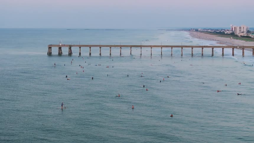 A long pier stretches into the ocean where several people are swimming and surfing near a shoreline with buildings